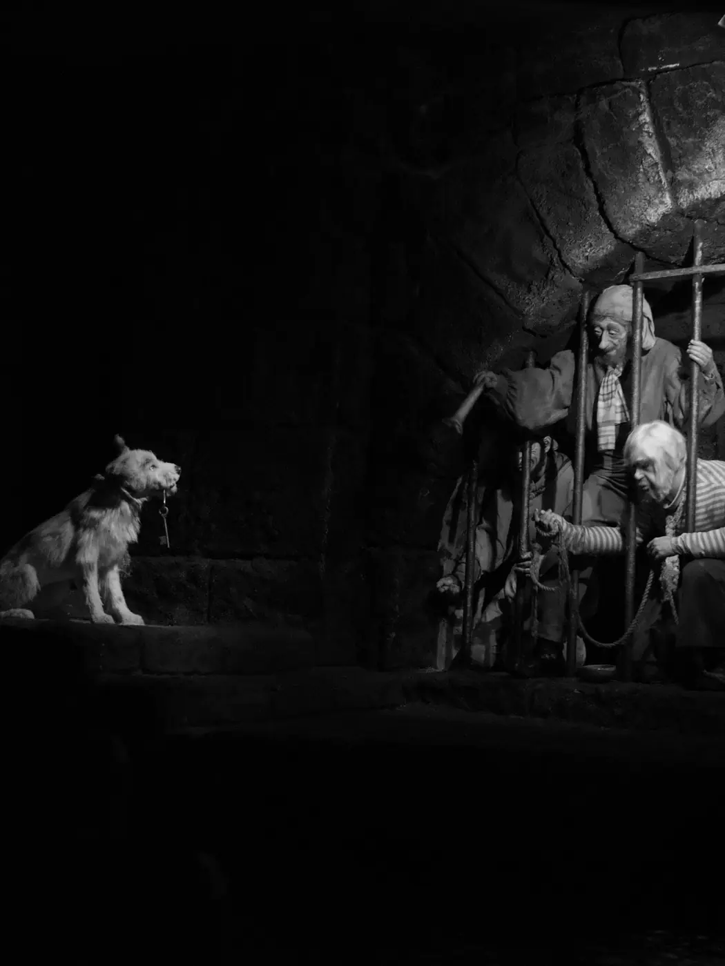 A dark ride scene in black and white: a dog holding a key in its mouth, sitting across from figures reaching through the bars of a cell